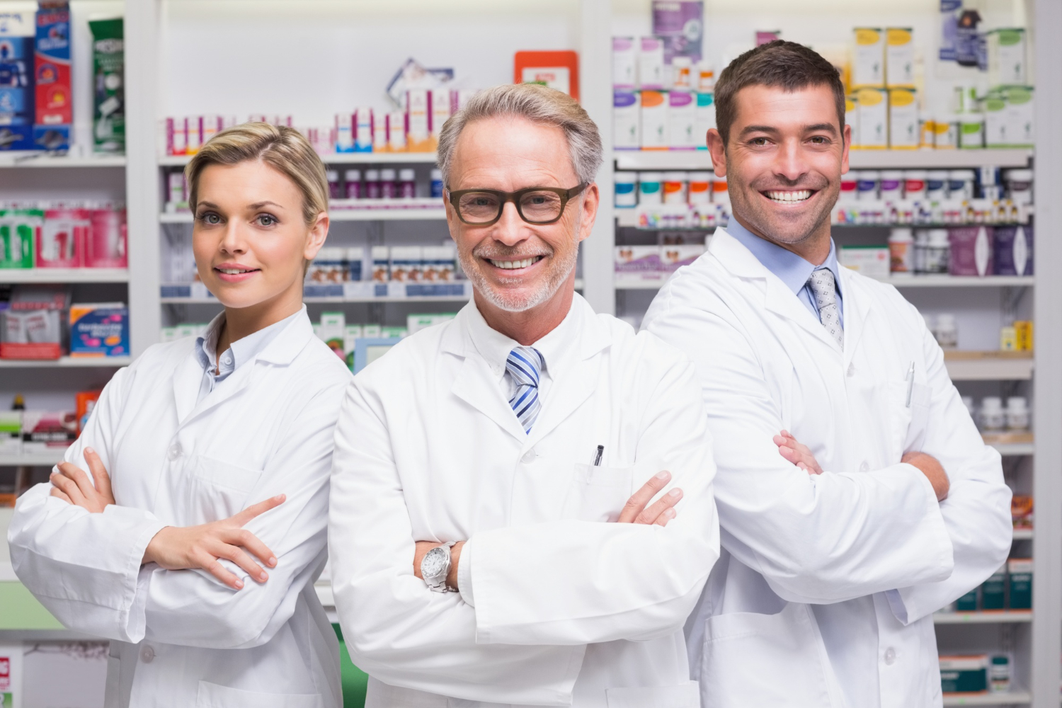 Three smiling pharmacists in white lab coats stand with arms crossed in a pharmacy.