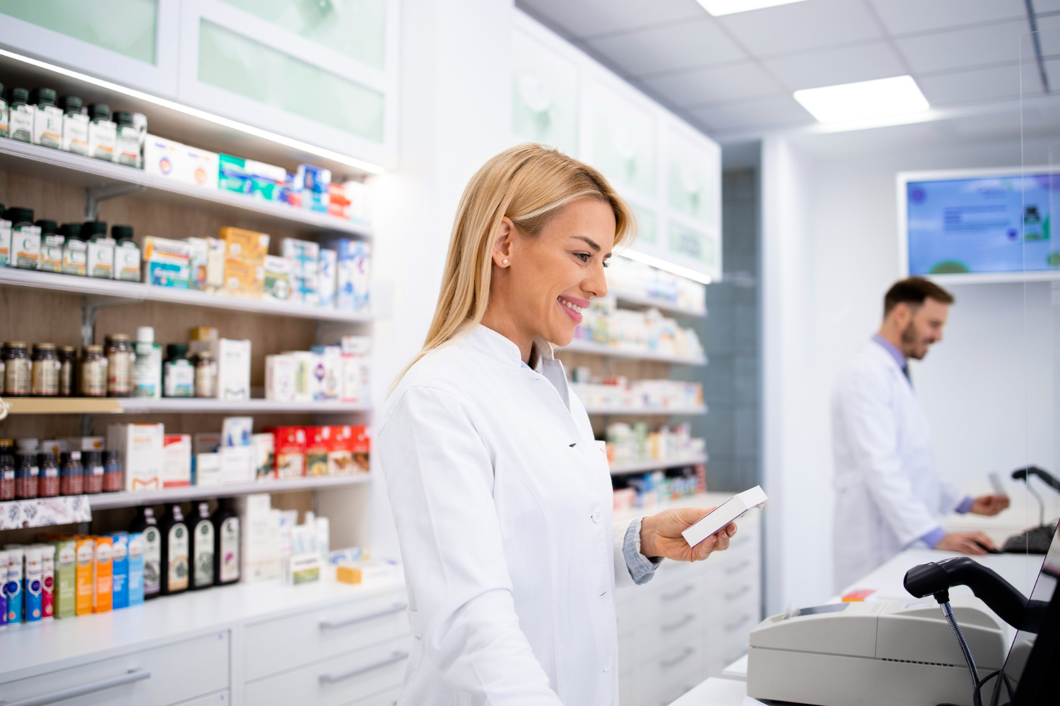 Smiling pharmacist in white coat holding medicine behind pharmacy counter, with shelves of products in the background.