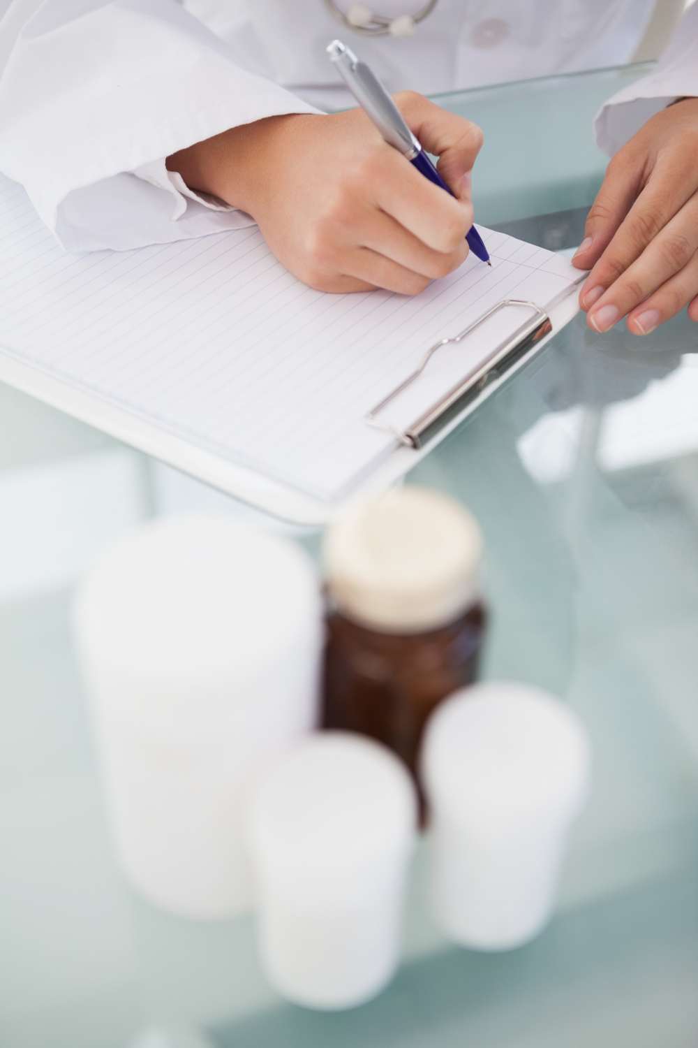 A doctor writing on a clipboard with medicine bottles on the table in the foreground.