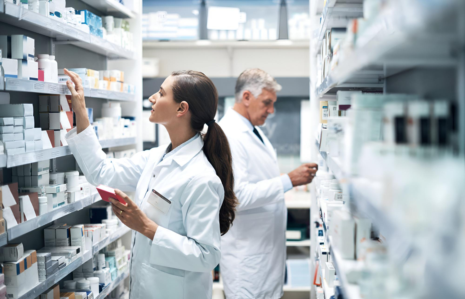 Two pharmacists in lab coats organize and check medications on shelves in a pharmacy storeroom.
