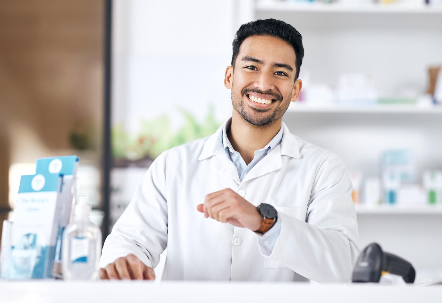 Smiling pharmacist in a white coat standing behind a counter in a pharmacy.
