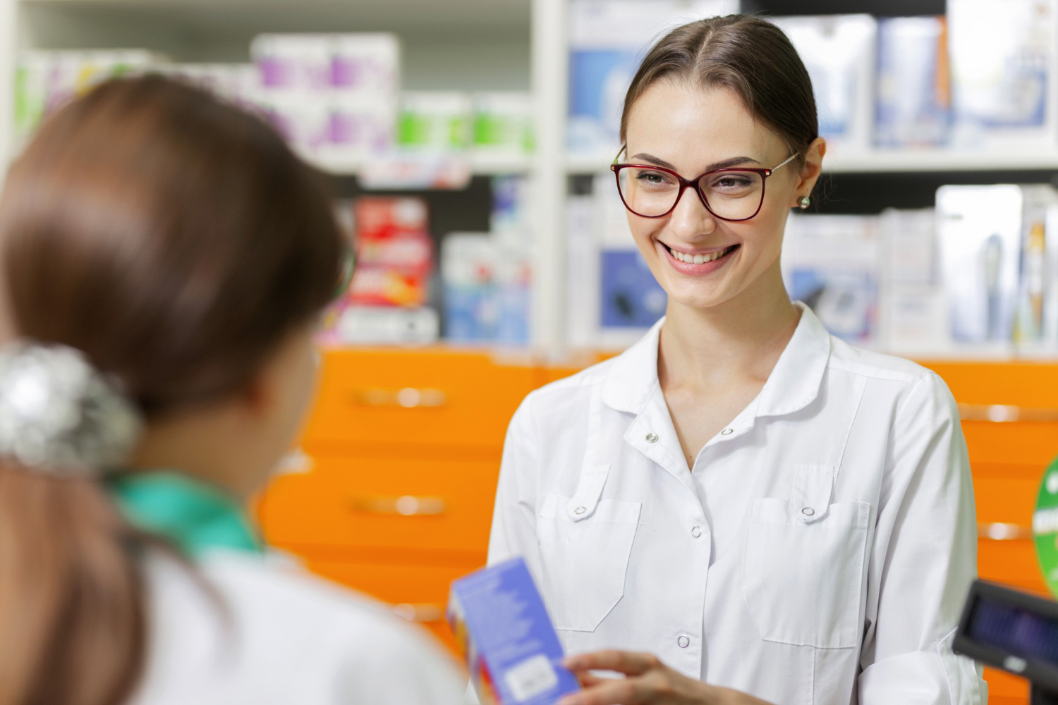 A smiling pharmacist assists a customer at a pharmacy counter, holding a blue box.