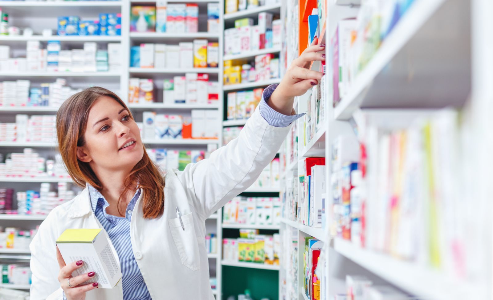 A pharmacist reaches for a box on a pharmacy shelf while holding another box in her hand.
