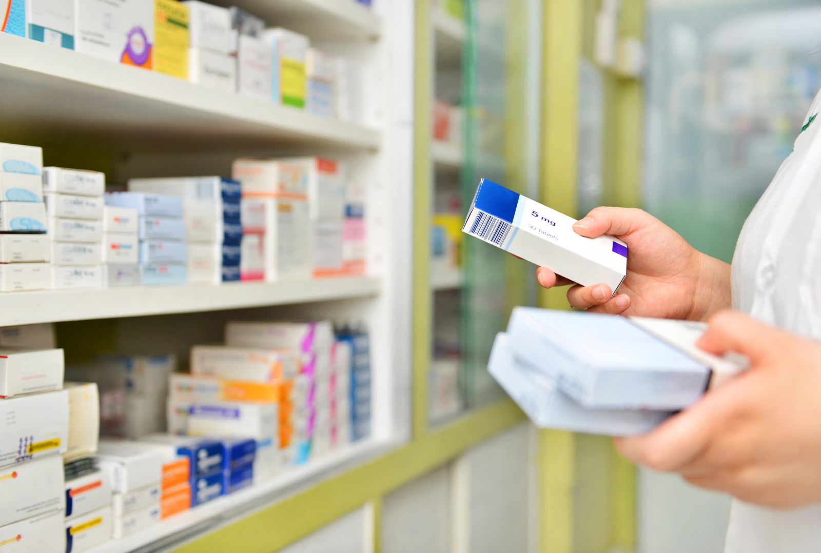 A pharmacist holds medicine boxes while standing in front of shelves filled with medications.