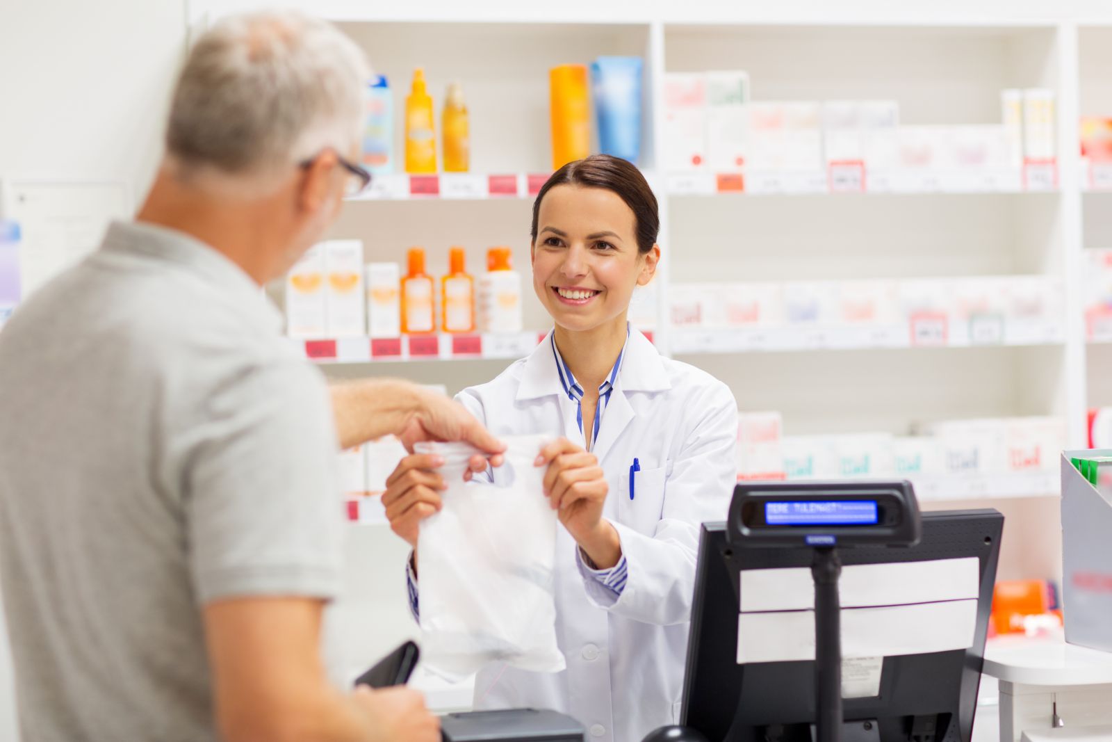 A pharmacist hands a prescription bag to a customer at a pharmacy counter, both smiling.