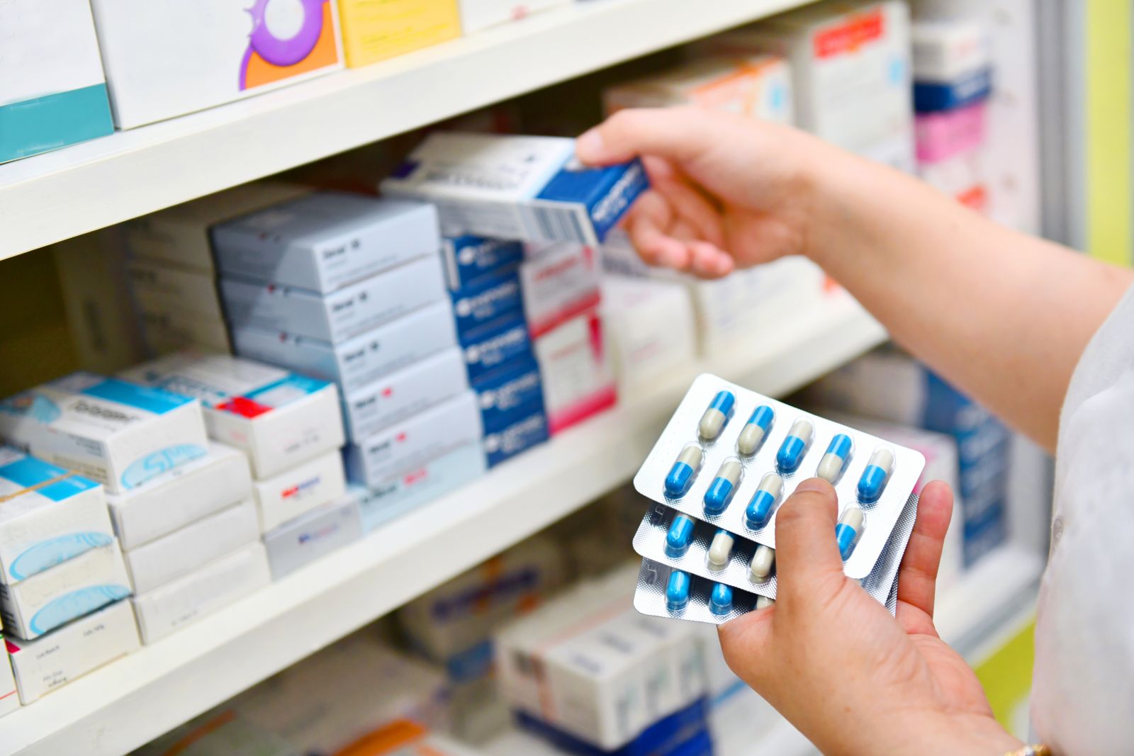 A person takes a box of medication from a pharmacy shelf while holding blister packs of capsules.