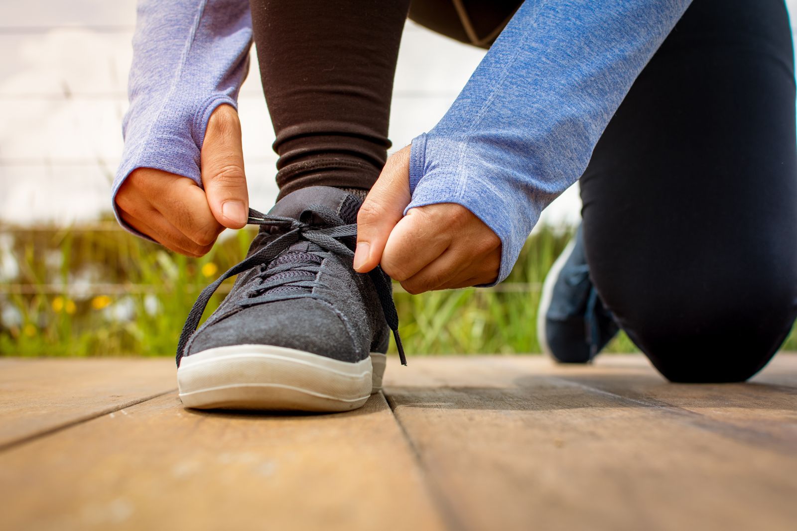 Person kneeling outdoors, tying the laces of a black sneaker on a wooden surface.
