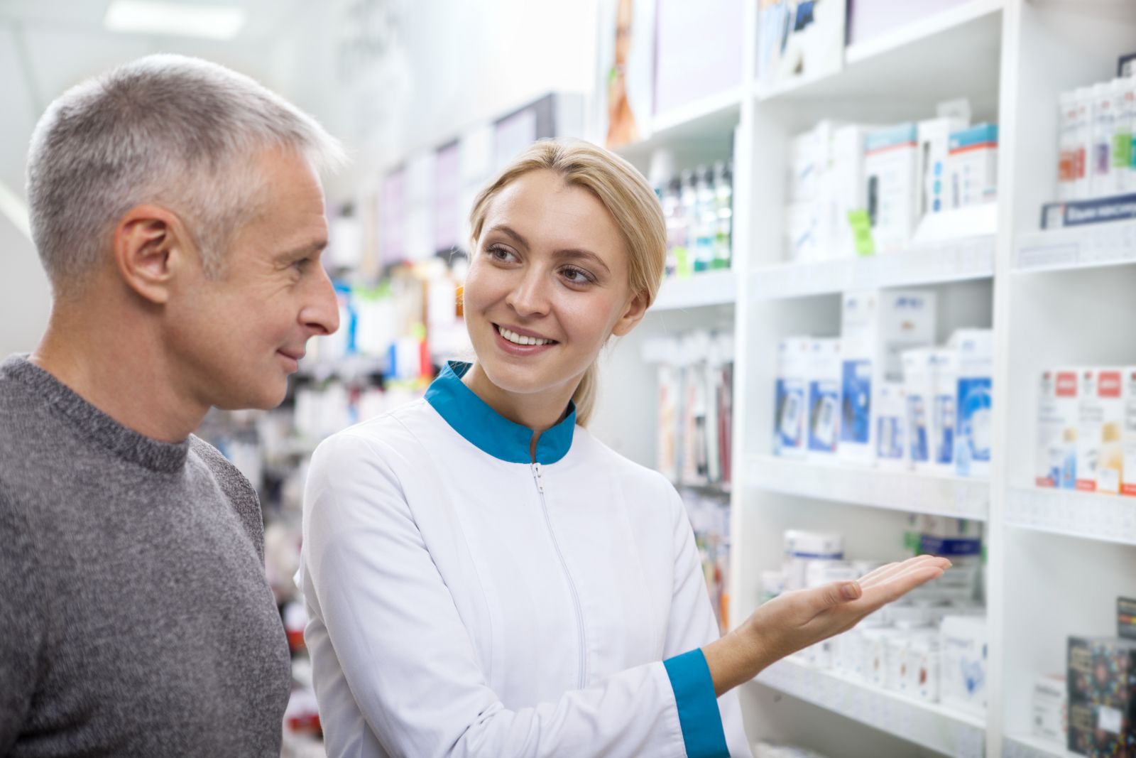 A pharmacist smiles and gestures to shelves of medicine while assisting a customer in a pharmacy.
