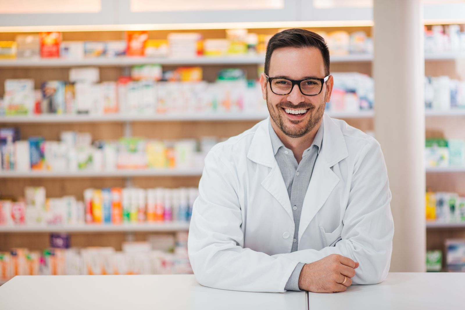 A smiling pharmacist in a white coat stands in a pharmacy with shelves of medicines behind him.
