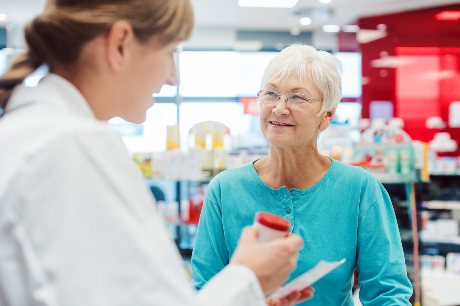 Smiling older woman talks to a pharmacist holding a prescription bottle in a pharmacy.