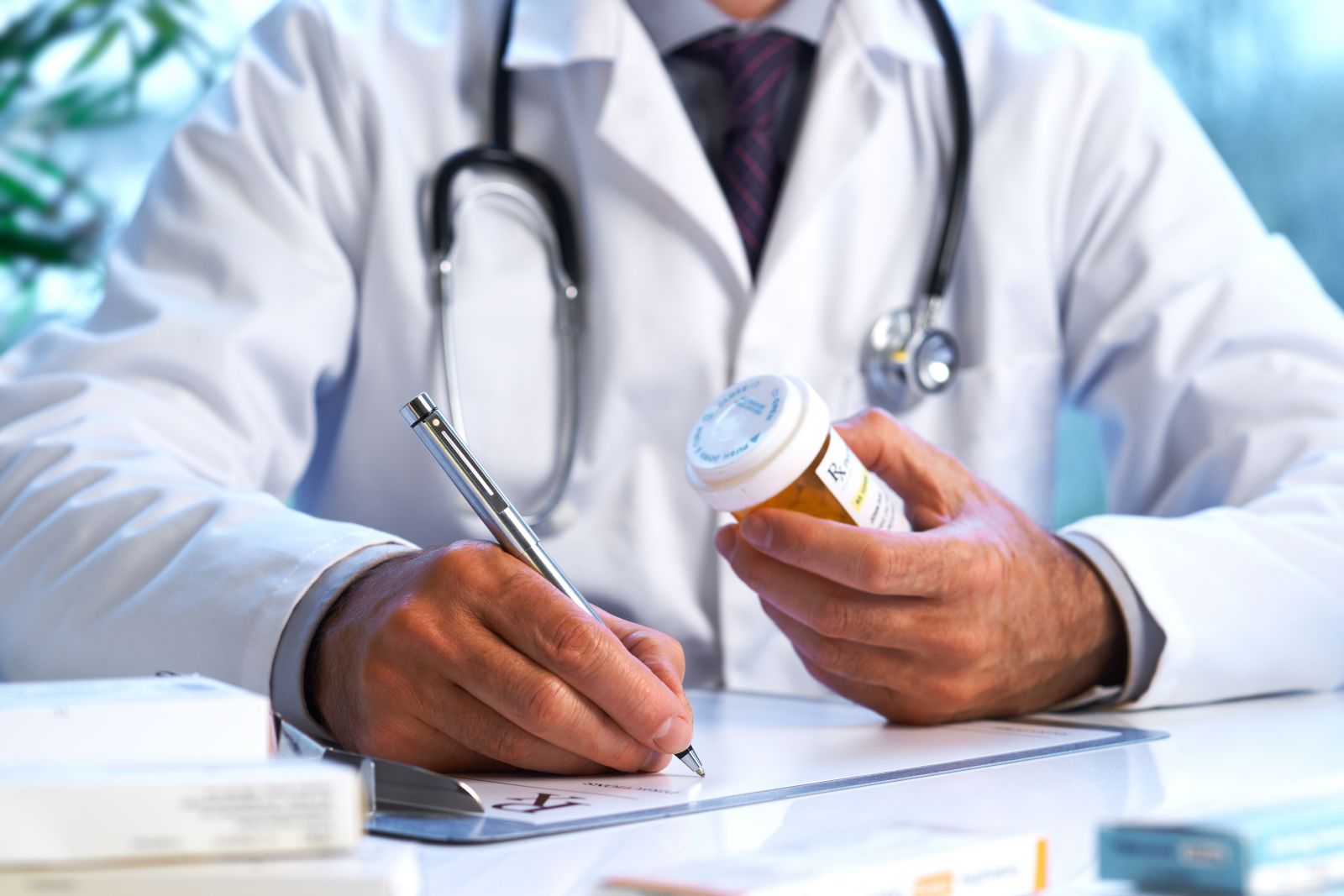 Doctor in white coat writes a prescription while holding a pill bottle at a desk with medical supplies.
