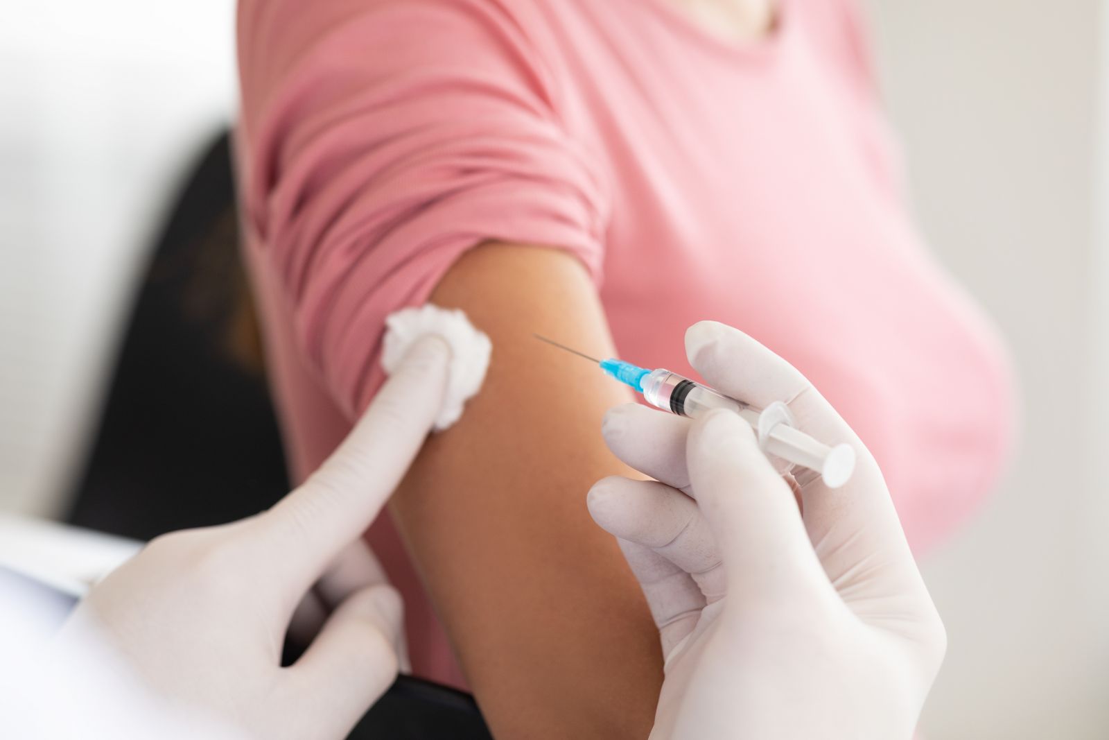 A person in a pink shirt receives an injection in their upper arm from a gloved healthcare worker.