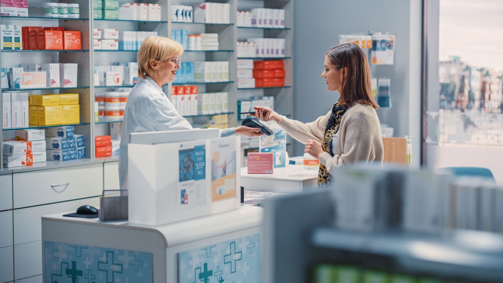 A woman pays a pharmacist at a modern pharmacy counter surrounded by shelves of medicine and products.