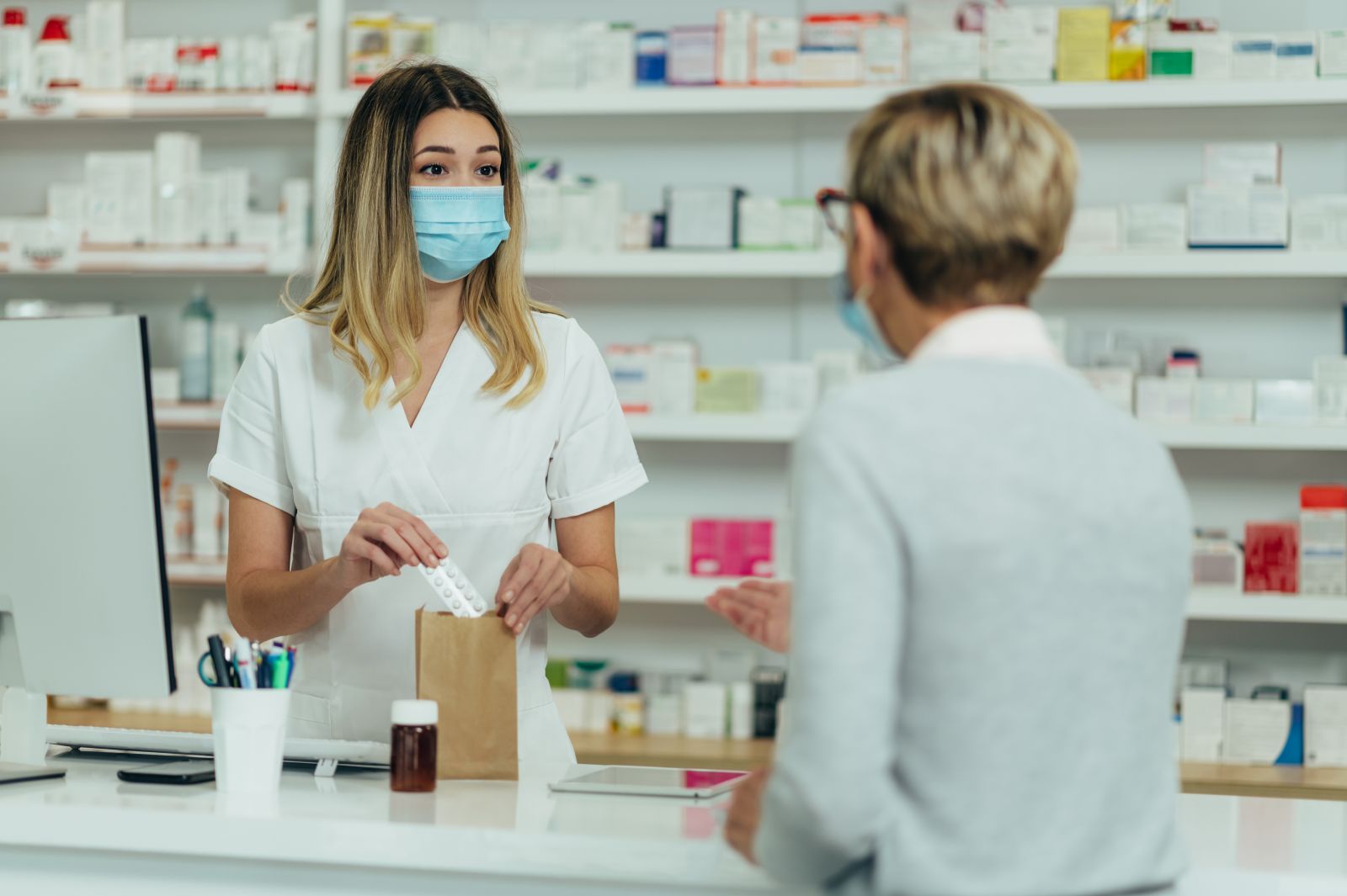 Pharmacist wearing a mask hands medicine in a paper bag to a masked customer at the pharmacy counter.