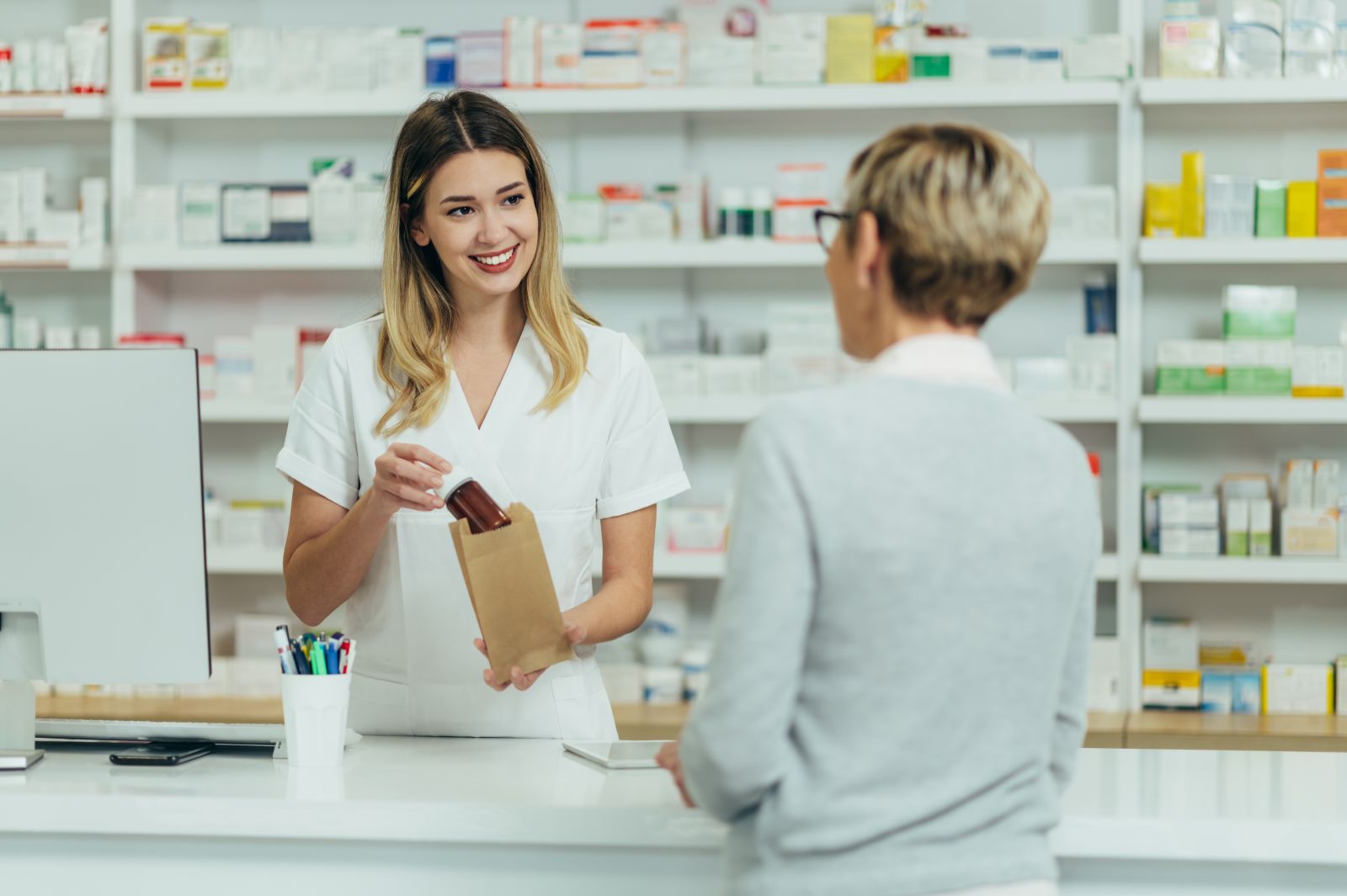 A pharmacist hands a prescription bottle in a paper bag to a customer at the pharmacy counter.