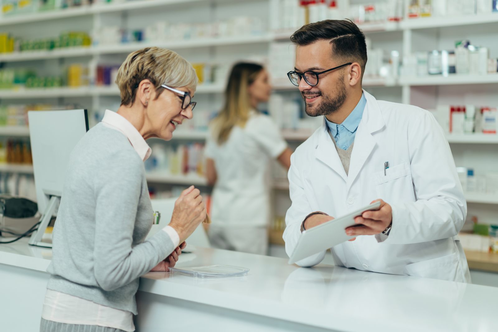 A pharmacist shows a tablet to a woman at a pharmacy counter while another worker is in the background.