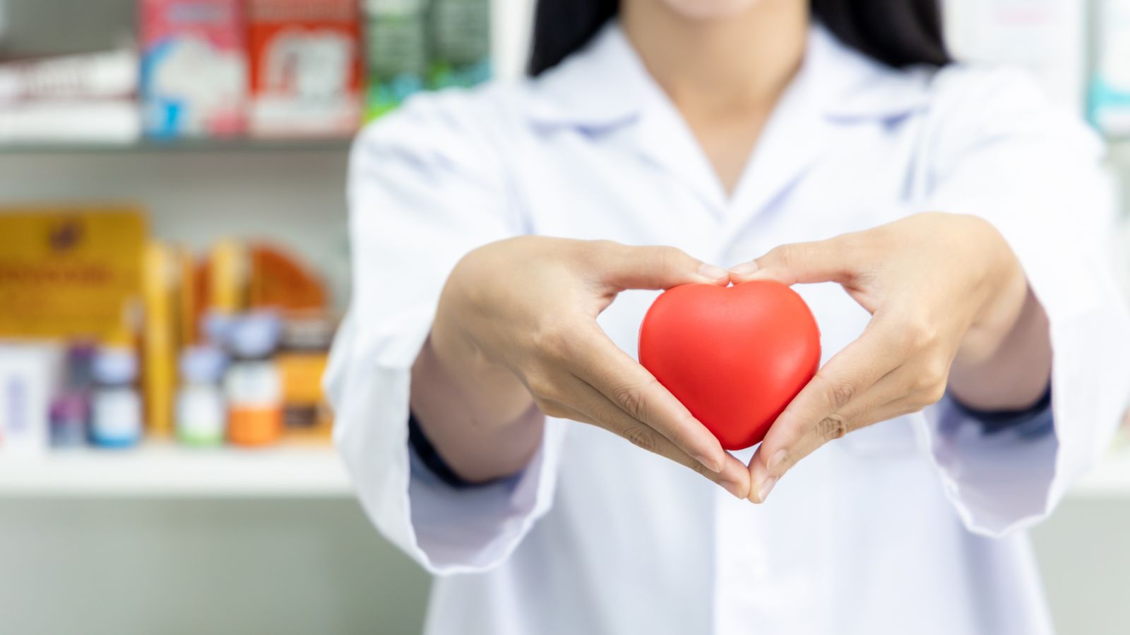 A person in a lab coat holds a red heart-shaped object in their hands in a pharmacy setting.