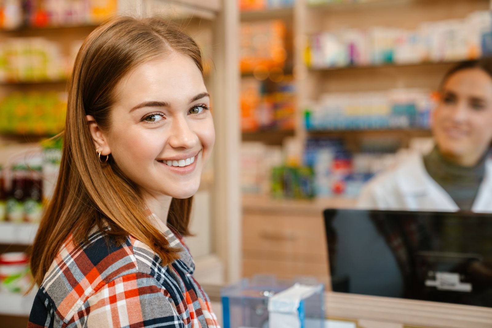 Young woman smiling at a pharmacy counter with a pharmacist in the background and shelves of products.