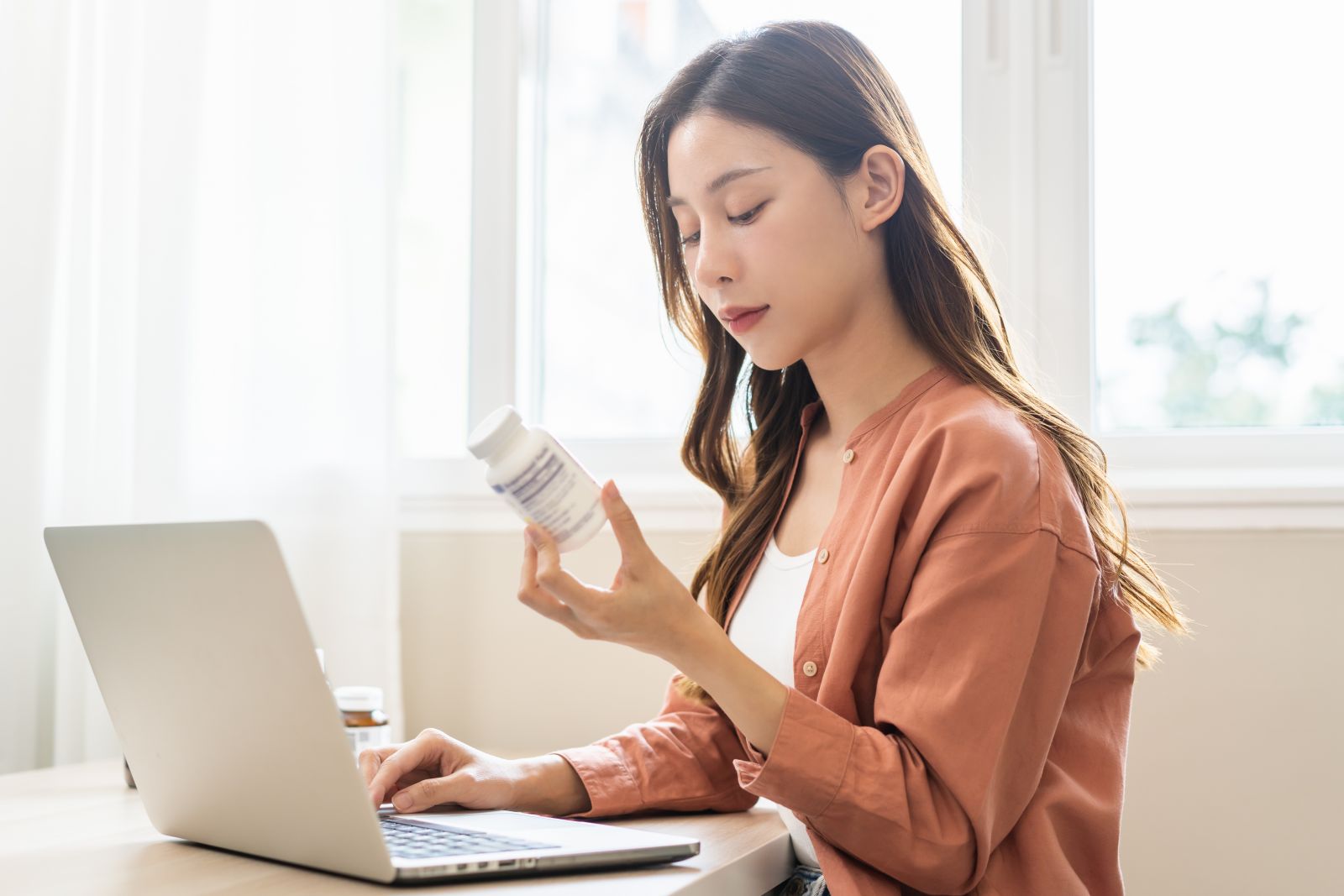 Woman sitting at a desk using a laptop and reading the label on a bottle of pills.