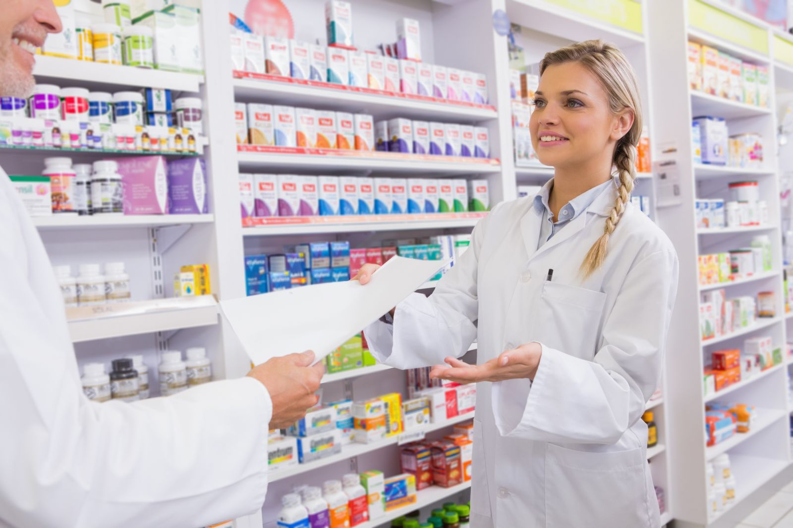 Pharmacist handing a prescription to a customer in a pharmacy with shelves of medicine in the background.
