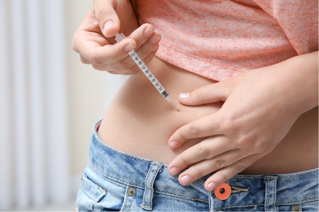 Person injecting a syringe into their abdomen, wearing a coral shirt and blue jeans.