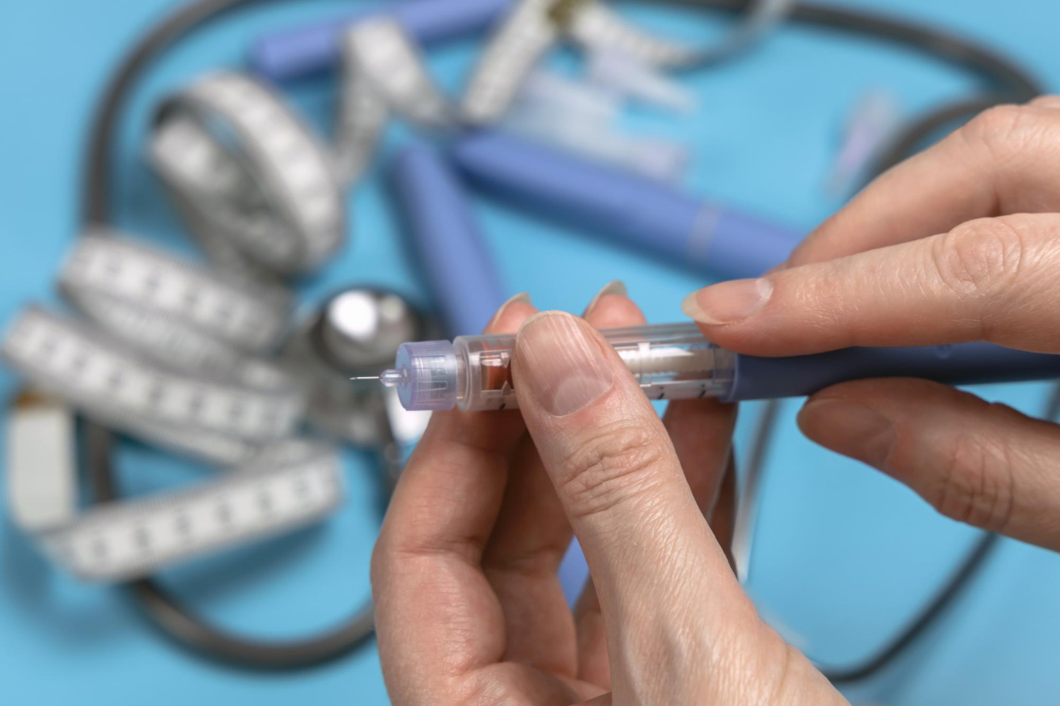Hands holding an insulin pen, with a tape measure and stethoscope in the background on a blue surface.