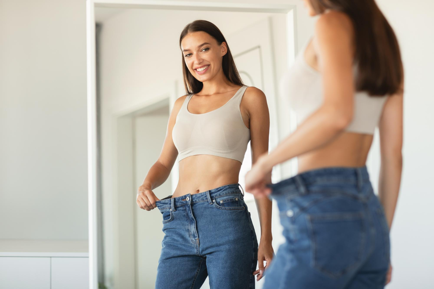 Woman smiling in front of a mirror, wearing loose jeans and holding the waistband to show weight loss.