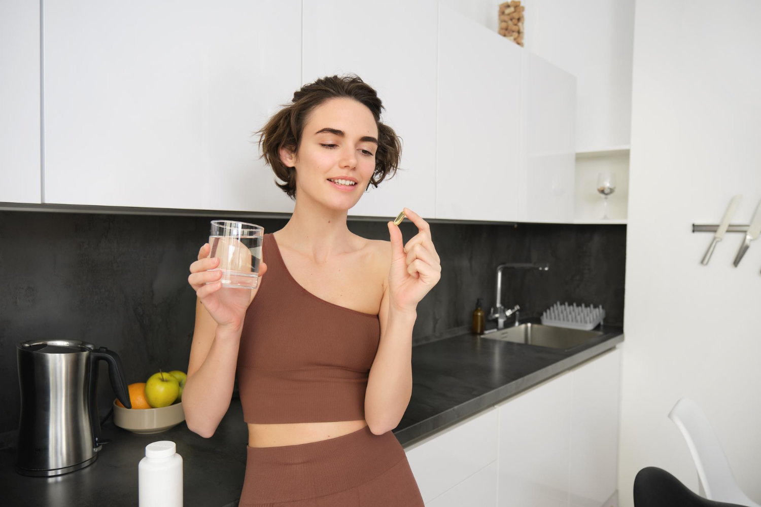 Woman in a brown outfit holding a glass of water and a pill in a modern white kitchen.