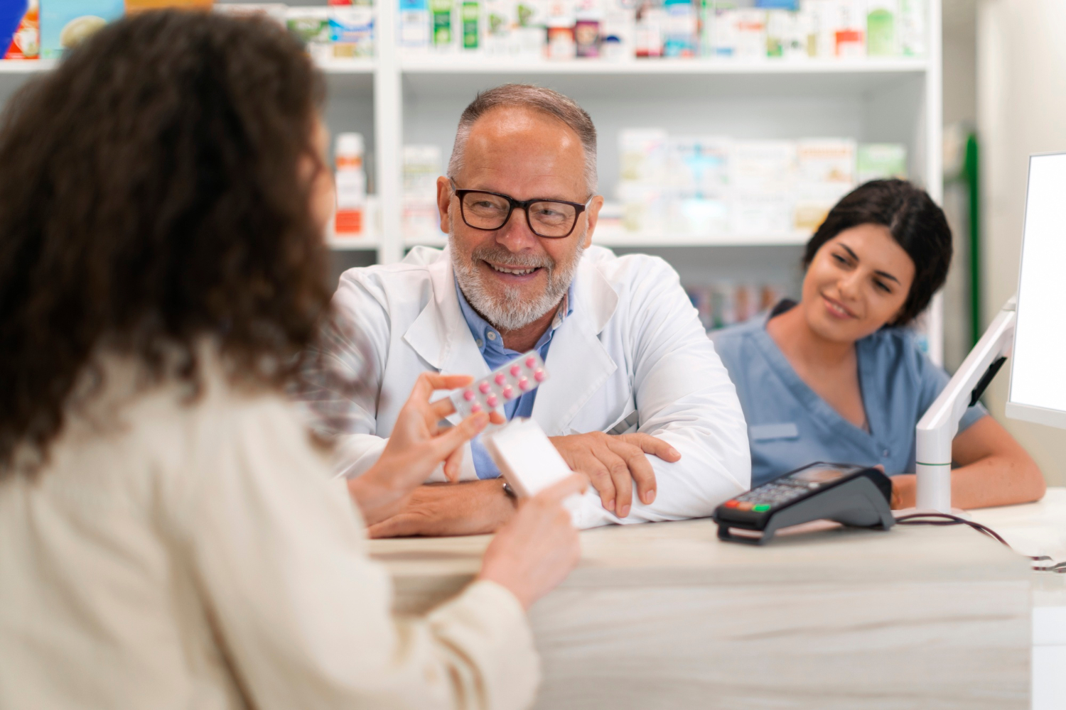 A pharmacist and assistant smile while helping a customer holding medication at the pharmacy counter.