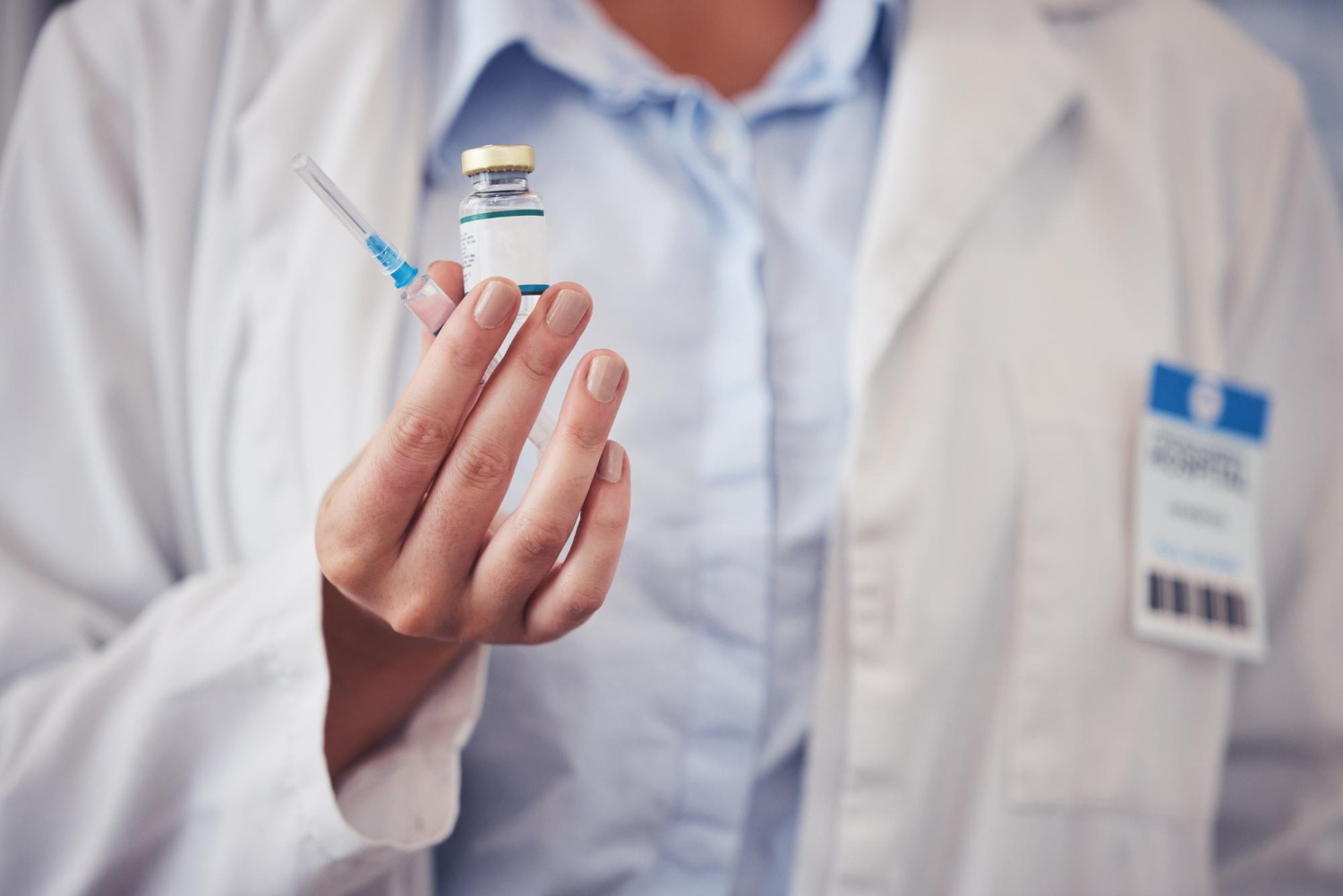A healthcare worker holds a syringe and a vial, wearing a white coat and name badge.