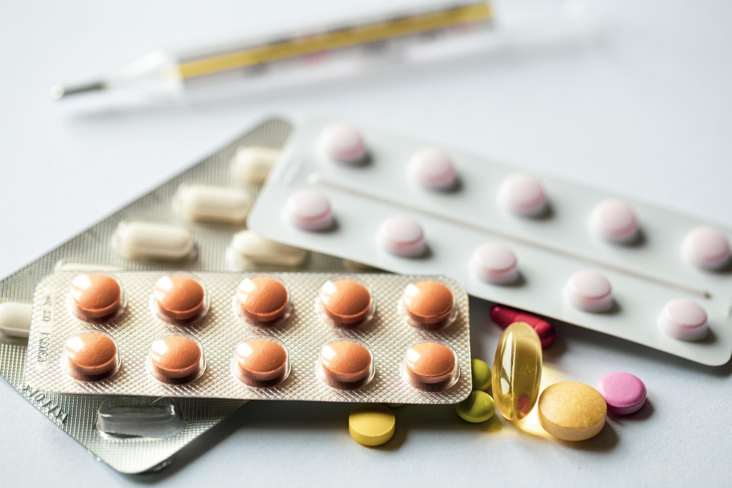 26625 Blister packs of assorted pills and tablets, with some loose pills and a thermometer on a white surface.